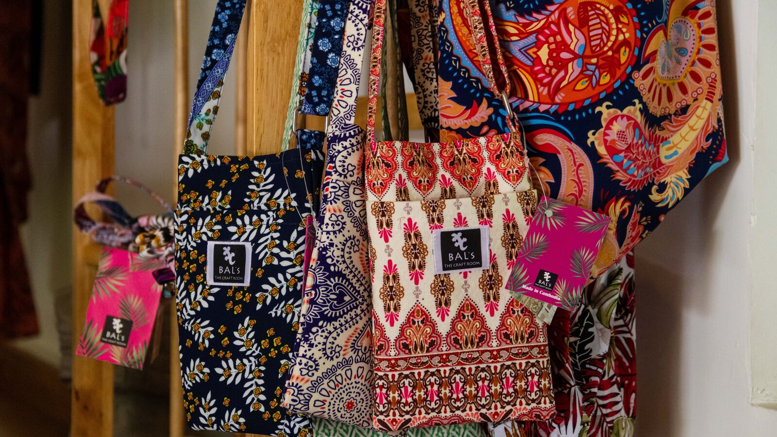 Collection of patterned fabric handbags made in Cambodia, hanging on a wooden display rack.