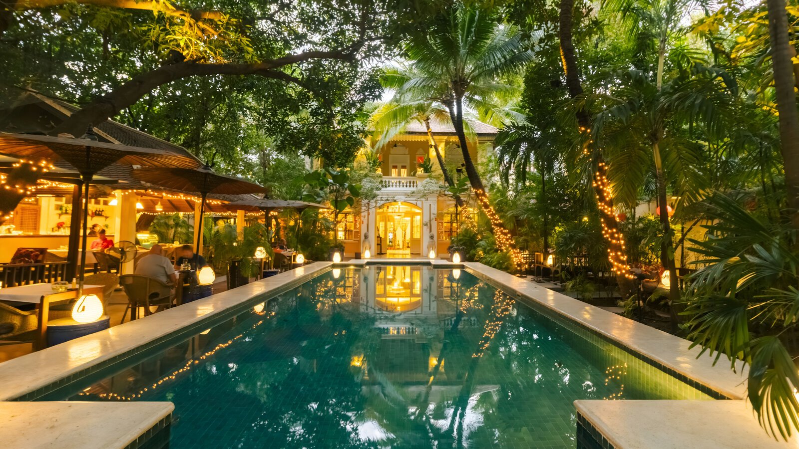Warm evening lights reflect across the pool toward the heritage villa surrounded by palms and garden.
