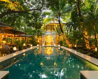 Warm evening lights reflect across the pool toward the heritage villa surrounded by palms and garden.