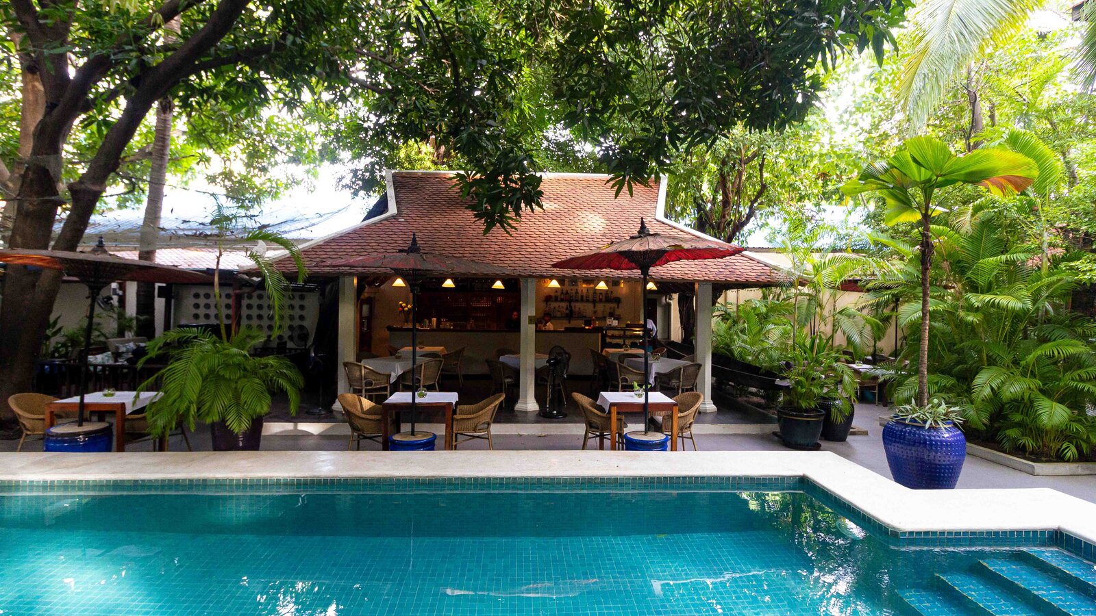 Open-air restaurant with traditional roof, surrounded by trees and plants by the pool.