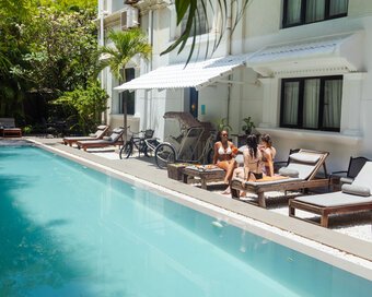 Three guests in swimwears chatting with drinks while seated on loungers beside the lap pool.