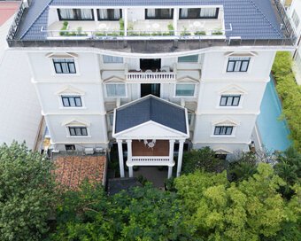 High drone view looking straight at the white facade and top-floor balconies of White Mansion Hotel in Phnom Penh, bordered by green trees and a lap pool.