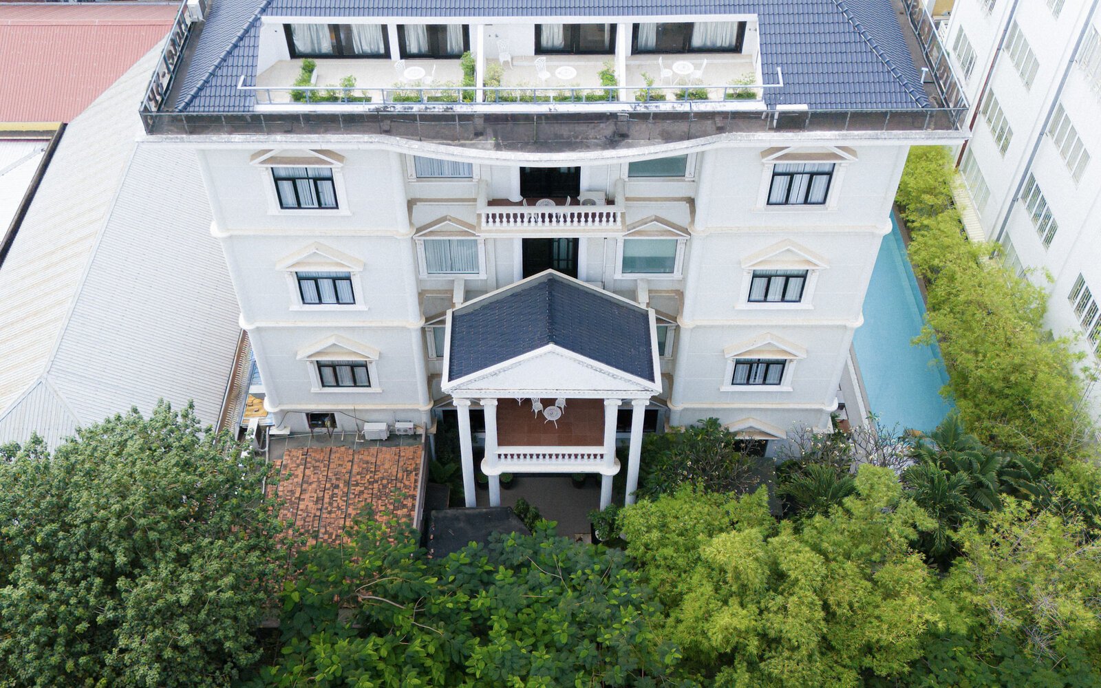 High drone view looking straight at the white facade and top-floor balconies of White Mansion Hotel in Phnom Penh, bordered by green trees and a lap pool.