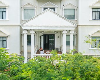 Guest sitting on a private white-column balcony at White Mansion, with green leafy branches, in Phnom Penh