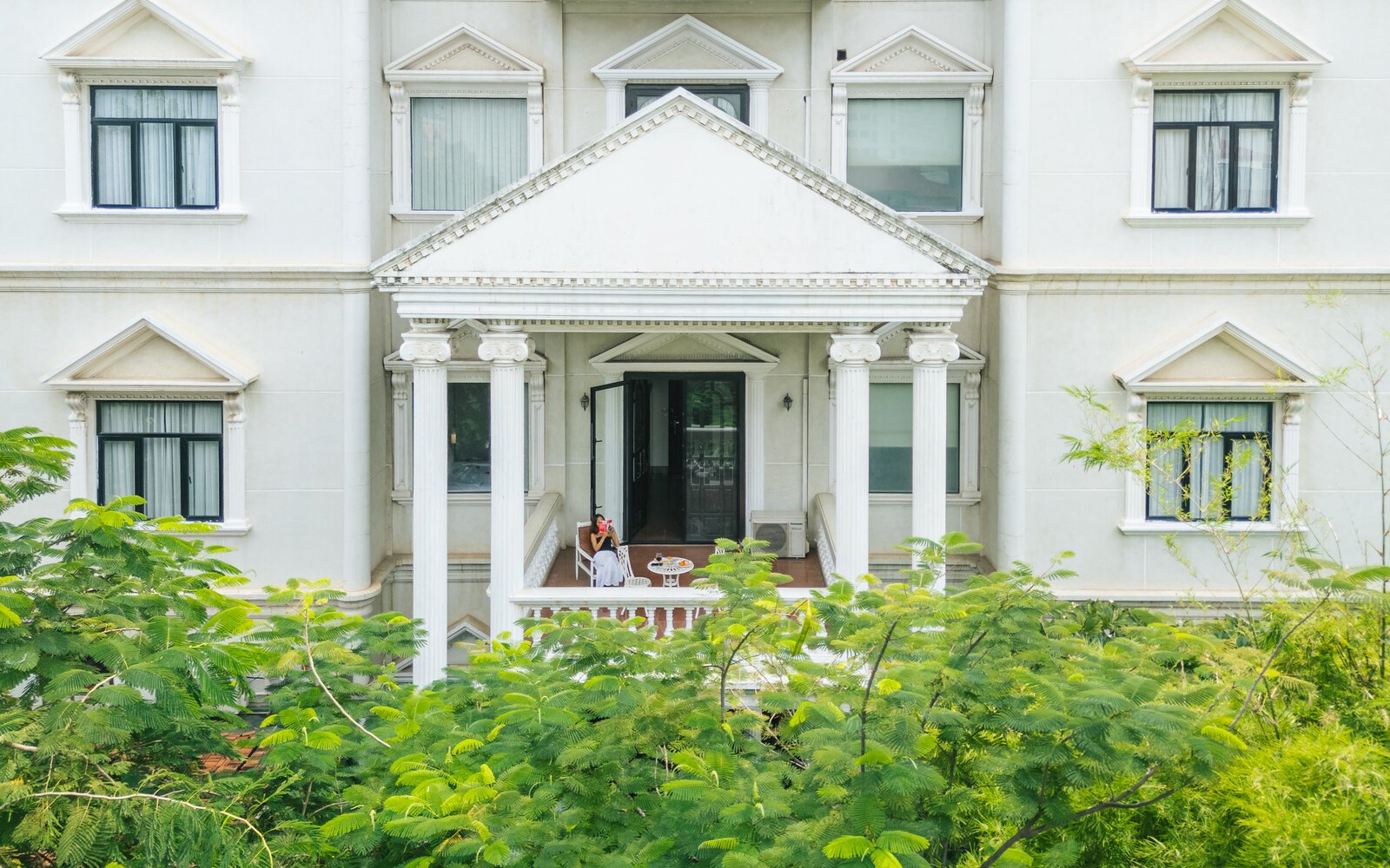 Guest sitting on a private white-column balcony at White Mansion, with green leafy branches, in Phnom Penh