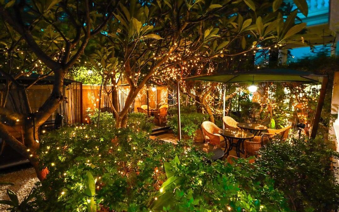 Outdoor café tables and woven chairs lit up by warm fairy lights in a leafy garden at Le Café, White Mansion, Phnom Penh