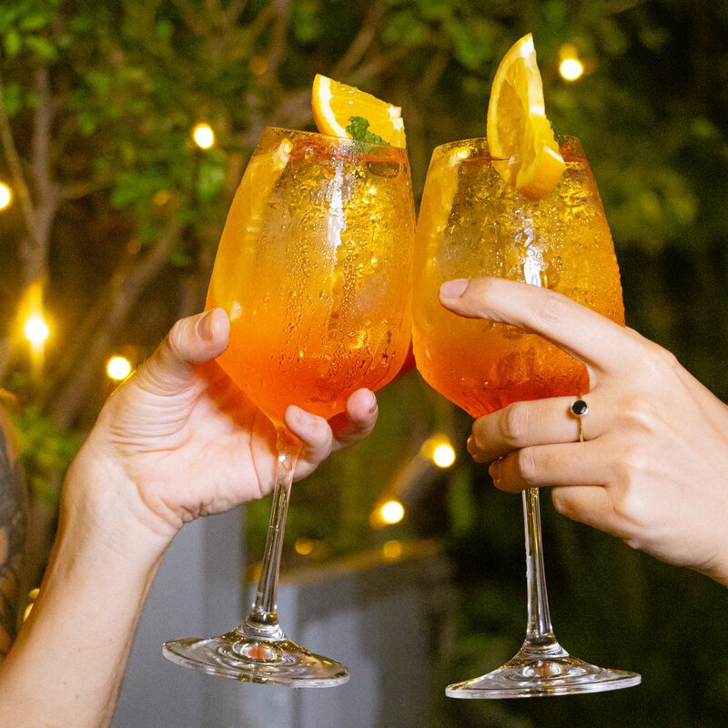 Close-up of two guests clinking orange spritz glasses garnished with citrus slices.