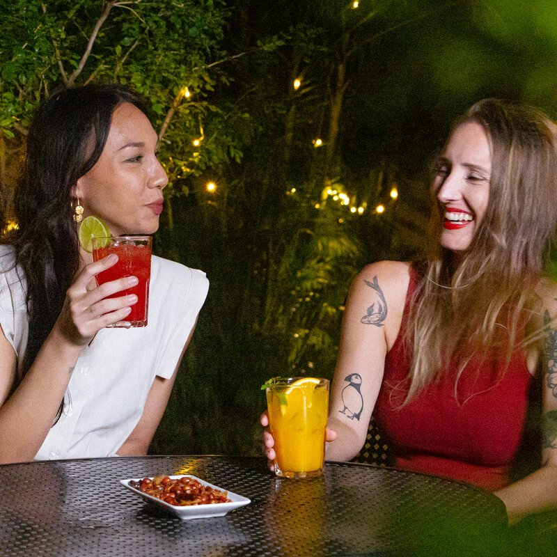 Two guests sharing laughs at Le Café garden with bright iced cocktails and appetizers.