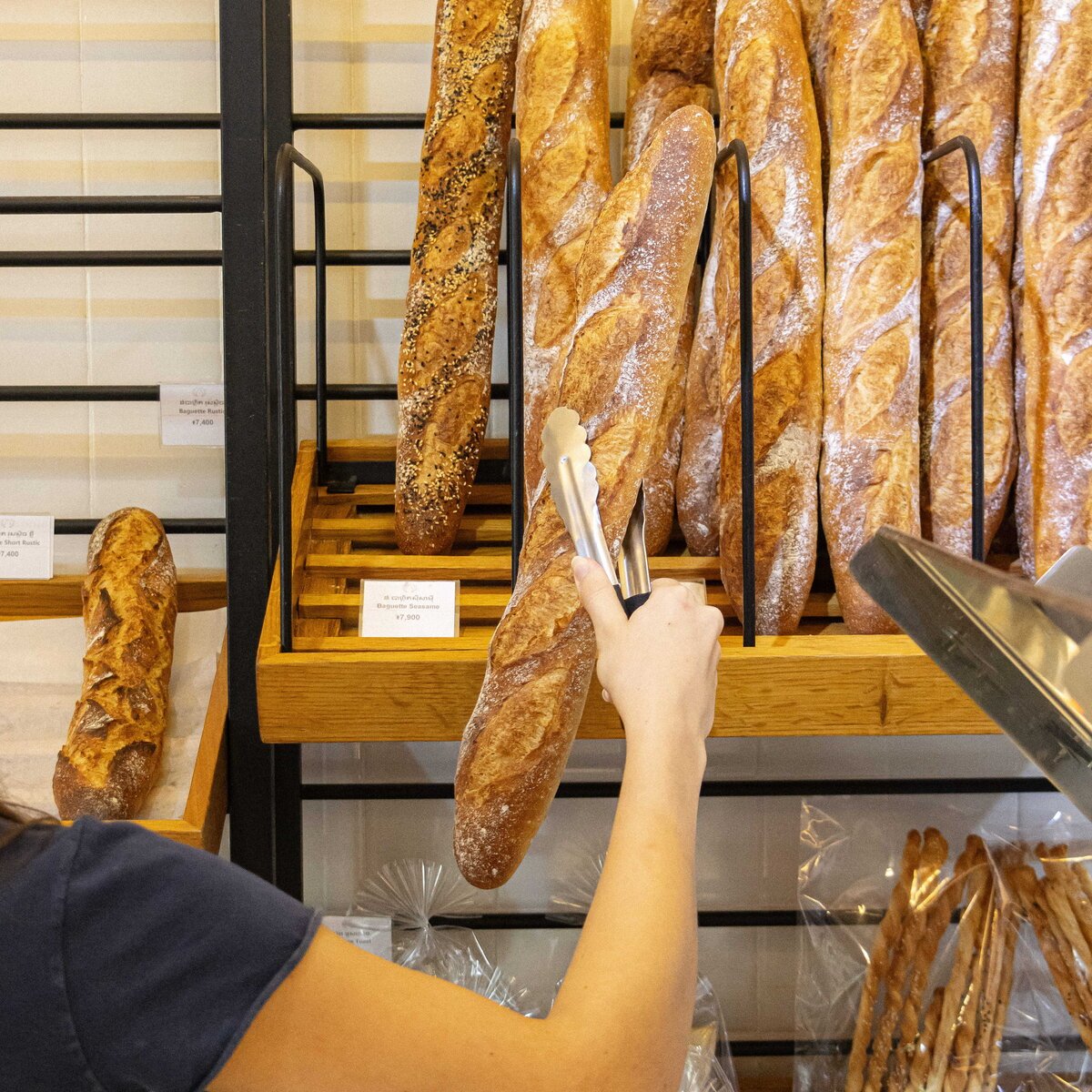 Hand holding tongs to pick a fresh baguette from a wooden display rack, Le Café at White Mansion Hotel, Phnom Penh