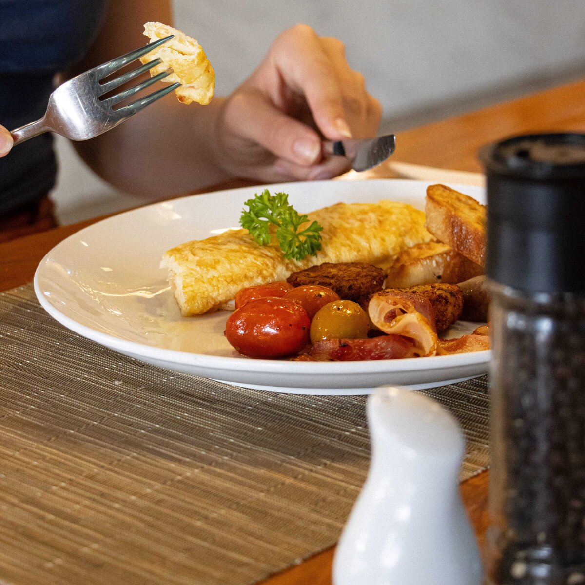 Customer taking a bite of an omelet served with grilled tomatoes, sausages, and toasted bread.