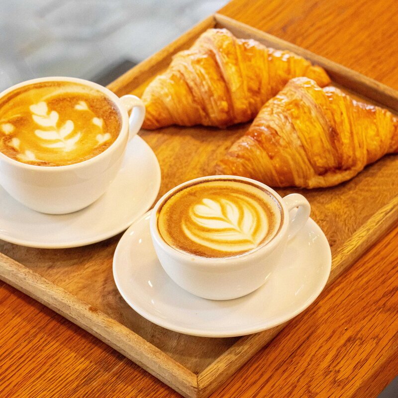 Two cups of hot latte with latte art served alongside two golden croissants on a wooden tray at Le Café, White Mansion Hotel, Phnom Penh.