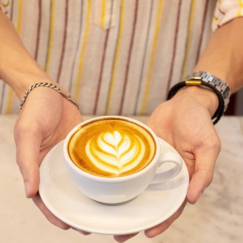Barista holding a white ceramic cup of hot cappuccino featuring detailed leaf latte art at Le Café, White Mansion Hotel, Phnom Penh.
